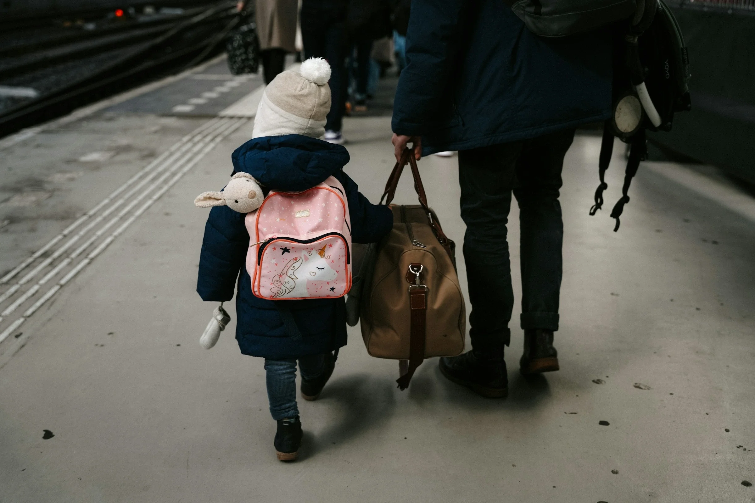 Niño pequeño de espaldas con gorro y mochila caminando junto a un adulto que lleva una bolsa de viaje